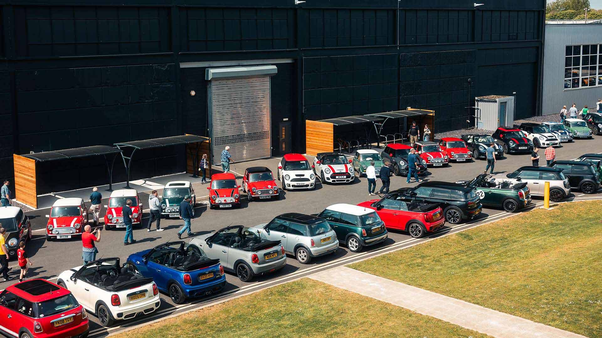A group of colorful Mini Coopers parked in rows outside a large black building under a partly cloudy sky, with people mingling around the cars.